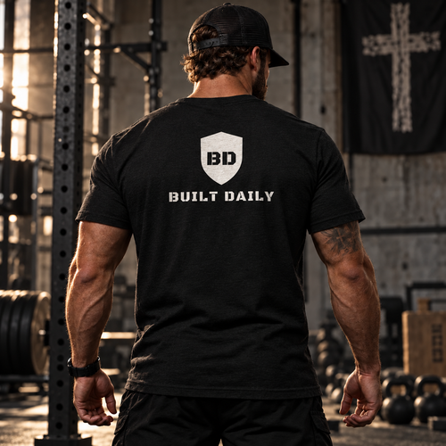 Man in a gym wearing the Built Daily Shield Tee, back view with the shield and BUILT DAILY