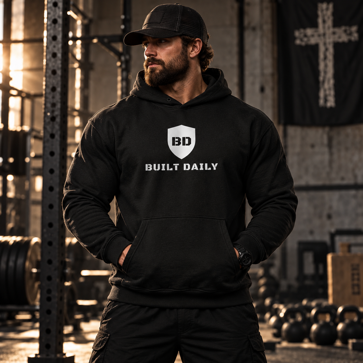 Man in a gym wearing the black Built Daily Shield Hoodie, front view with the shield and BUILT DAILY